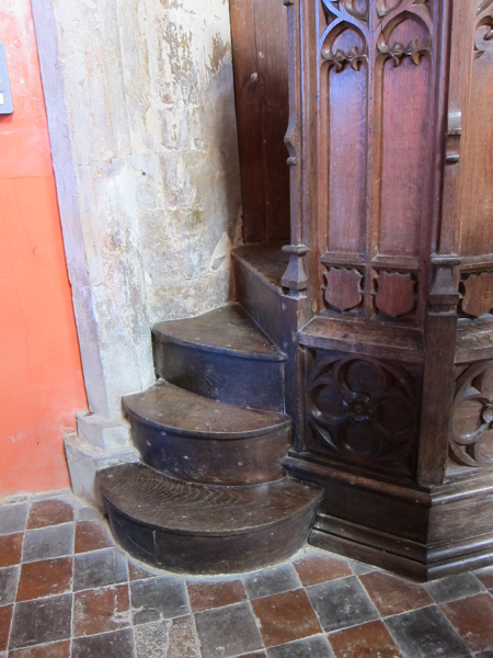 Colour photograph with a set of three wooden steps leading to a wooden pulpit, with a brown and grey tiled floor